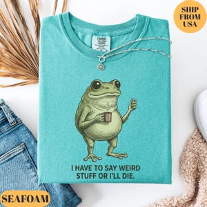 K&ouml;nnte beinhalten: T&uuml;rkisgr&uuml;nes T-Shirt mit einer Grafik einer Froschfigur, die eine Tasse h&auml;lt, und dem Text "I HAVE TO SAY WEIRD STUFF OR I'LL DIE." Eine silberne Halskette mit Sonnenanh&auml;nger befindet sich auf dem Shirt. Das Shirt ist aus Comfort Colors.