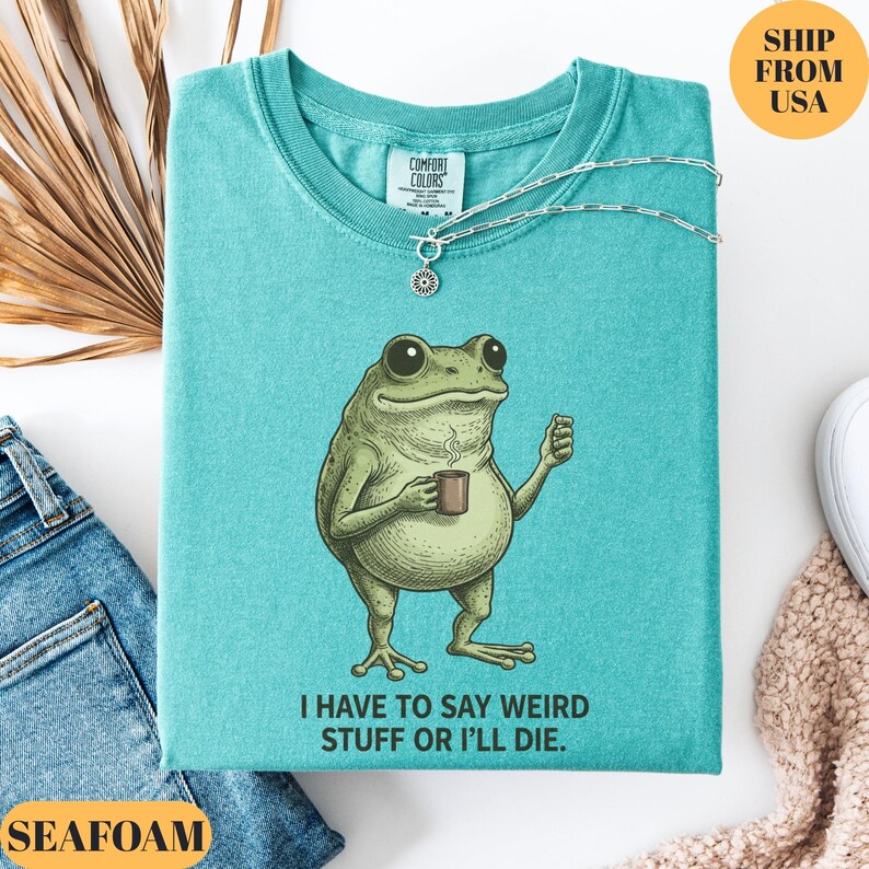K&ouml;nnte beinhalten: T&uuml;rkisgr&uuml;nes T-Shirt mit einer Grafik einer Froschfigur, die eine Tasse h&auml;lt, und dem Text "I HAVE TO SAY WEIRD STUFF OR I'LL DIE." Eine silberne Halskette mit Sonnenanh&auml;nger befindet sich auf dem Shirt. Das Shirt ist aus Comfort Colors.