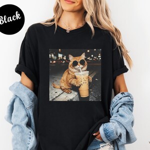 May include: Black t-shirt featuring a graphic of a cat wearing sunglasses and holding an iced coffee. The cat is orange with black sunglasses. The text "Black" is in a black circle in the upper left corner.