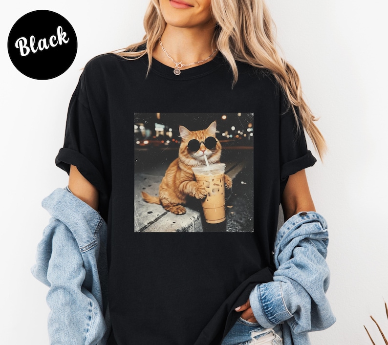 May include: Black t-shirt featuring a graphic of a cat wearing sunglasses and holding an iced coffee. The cat is orange with black sunglasses. The text "Black" is in a black circle in the upper left corner.