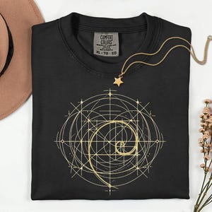 Könnte beinhalten: Schwarzes T-Shirt mit einem goldenen geometrischen Design mit einer Spirale und sich kreuzenden Linien. Eine goldene Sternanhänger-Halskette liegt auf dem Shirt. Das Etikett lautet "COMFORT COLORS XL-TG-EG".