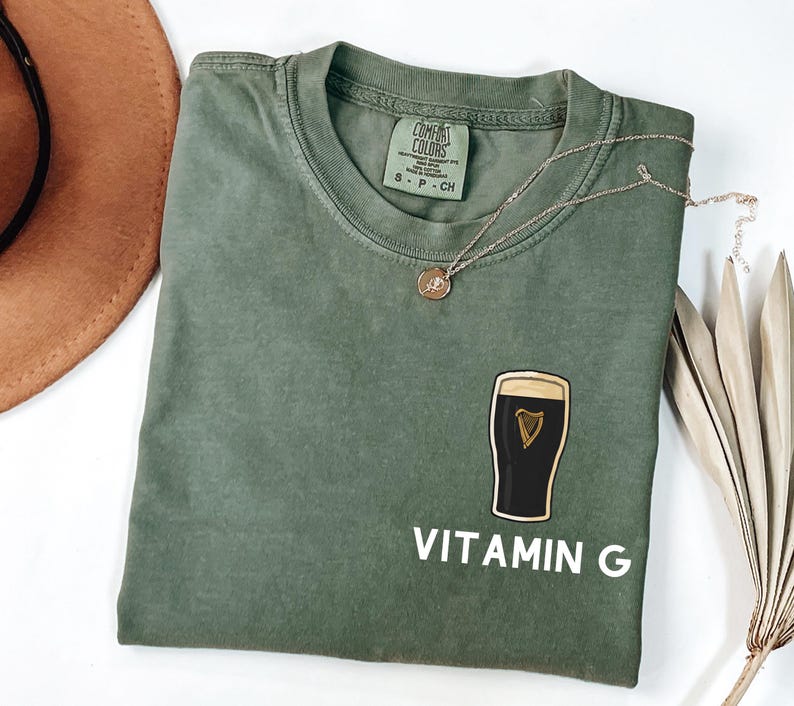 May include: An olive green t-shirt with a graphic of a pint of stout and the words "VITAMIN G" in white. A gold necklace with a small pendant is draped on the shirt. The shirt is folded, and a brown hat is partially visible.
