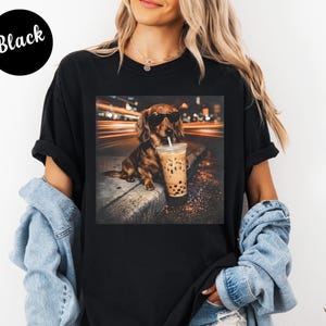 Puede incluir: Camiseta negra con un gr&aacute;fico de un perro salchicha con gafas de sol, sosteniendo un bubble tea. El perro es marr&oacute;n y canela, con una bebida en un vaso de pl&aacute;stico transparente. El fondo muestra luces borrosas.