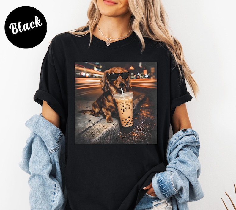 Puede incluir: Camiseta negra con un gr&aacute;fico de un perro salchicha con gafas de sol, sosteniendo un bubble tea. El perro es marr&oacute;n y canela, con una bebida en un vaso de pl&aacute;stico transparente. El fondo muestra luces borrosas.