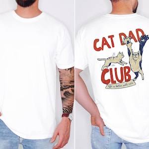 May include: White t-shirt with a graphic on the back that says "CAT DAD CLUB" in red and beige. The graphic features two cats, one jumping and the other reaching up. The text "life is better with cats" is below the word "CLUB".