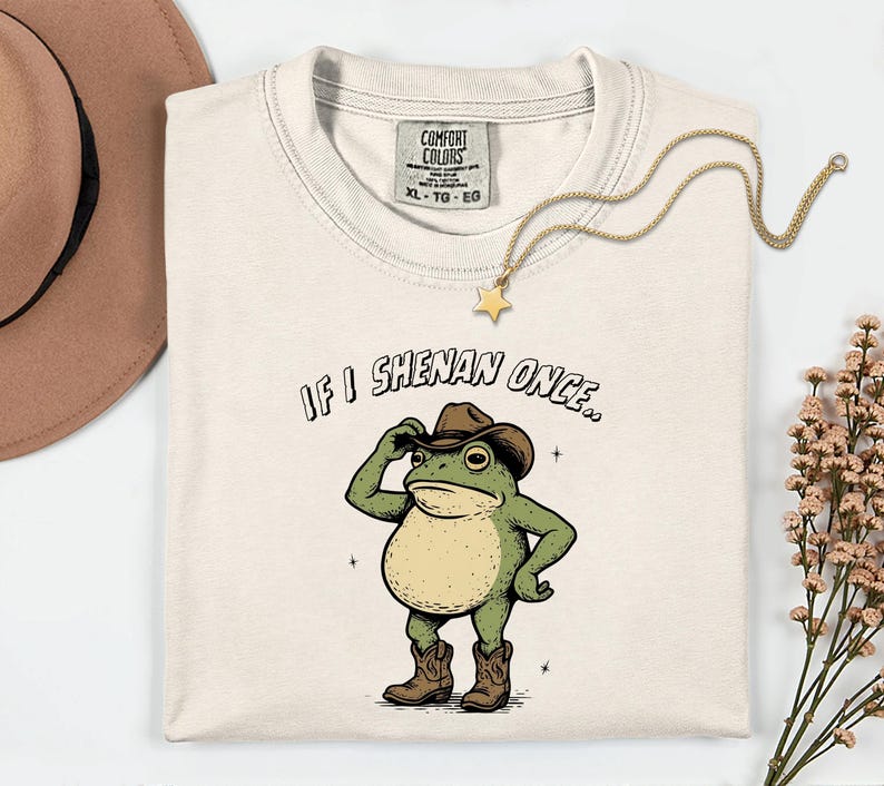 May include: Off-white t-shirt with a cartoon frog in a cowboy hat and boots. The frog is tipping its hat, with the text "IF I SHENAN ONCE.." above it. A gold necklace with a star pendant is on the shirt.