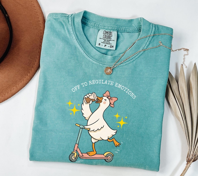 May include: Teal Comfort Colors t-shirt with a cartoon goose riding a pink scooter and drinking coffee. The text "Off to Regulate Emotions" is above the goose. A gold necklace is draped across the shirt.