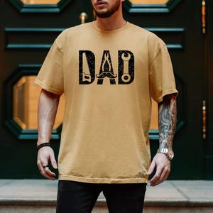 May include: Mustard yellow t-shirt featuring the word "DAD" in black, block letters. The letters are designed with tools and a moustache. The shirt is short-sleeved and has a crew neck.