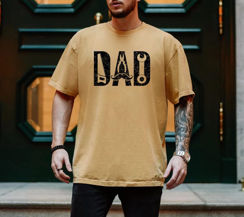 May include: Mustard yellow t-shirt featuring the word "DAD" in black, block letters. The letters are designed with tools and a moustache. The shirt is short-sleeved and has a crew neck.