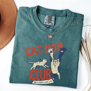 May include: Teal Comfort Colors t-shirt with the words "CAT MOM CLUB" in red, featuring cat illustrations. A gold necklace with a pendant rests on the shirt. The phrase "life is better with cats" is printed at the bottom.