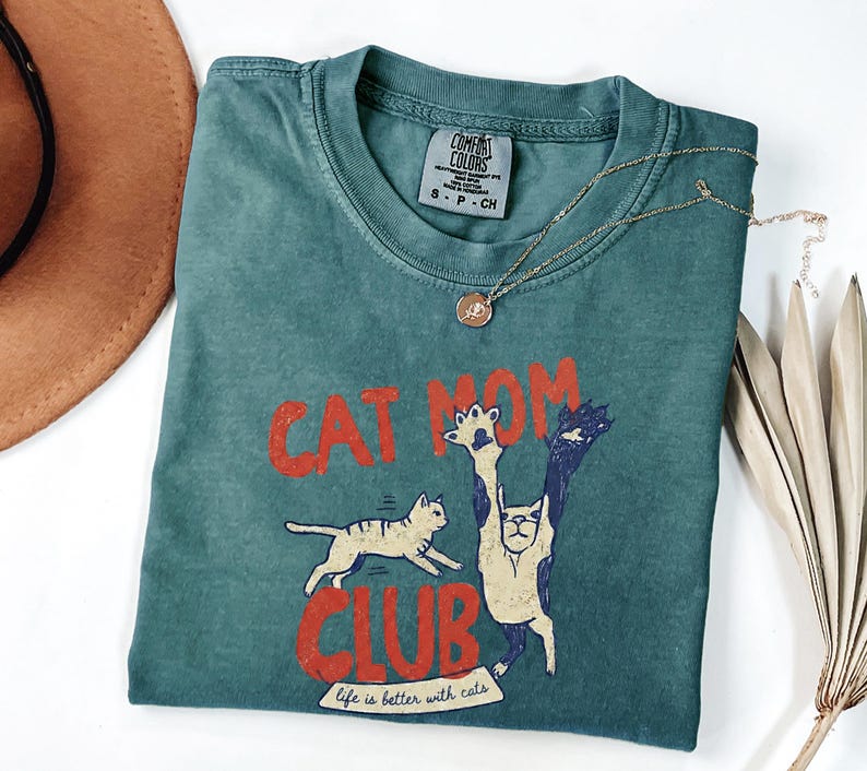 May include: Teal Comfort Colors t-shirt with the words "CAT MOM CLUB" in red, featuring cat illustrations. A gold necklace with a pendant rests on the shirt. The phrase "life is better with cats" is printed at the bottom.