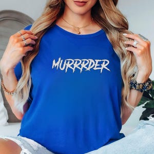 May include: A royal blue t-shirt with the word "MURRRDER" in a white, jagged font. The shirt is a solid color and has a crew neck. The text is centered on the front of the shirt.