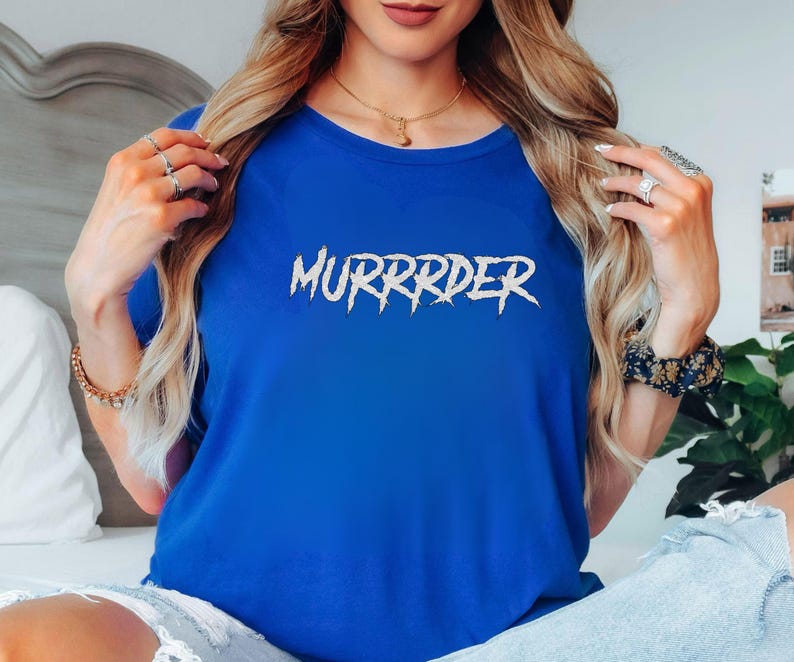 May include: A royal blue t-shirt with the word "MURRRDER" in a white, jagged font. The shirt is a solid color and has a crew neck. The text is centered on the front of the shirt.