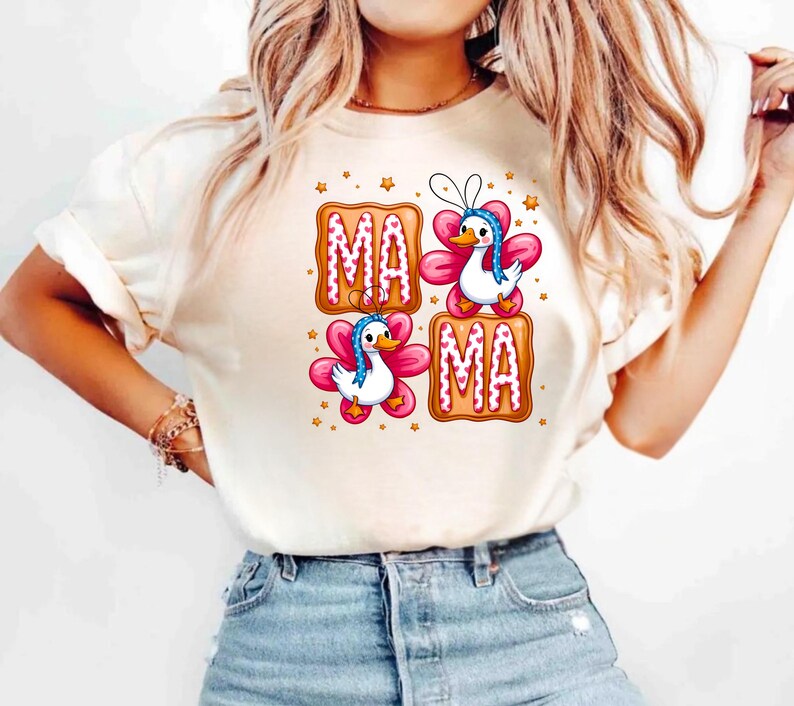 May include: Cream-colored t-shirt with a graphic design featuring the word "MAMA" in a gingerbread cookie style. The design includes cartoon ducks with blue bonnets and pink flower wings. Small gold stars are scattered around the design.