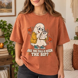 May include: A rust-coloured short-sleeve t-shirt featuring a graphic of a ghost wearing glasses and a yellow cardigan, holding a clipboard labelled BIP. The text reads Are You Fall-o-ween The Bip? with orange maple leaves surrounding the ghost.