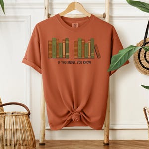 May include: Rust-colored t-shirt featuring a graphic of a bookshelf with green, brown, and tan books. The text "IF YOU KNOW, YOU KNOW" is printed below the bookshelf. The shirt is hanging on a wooden rack.