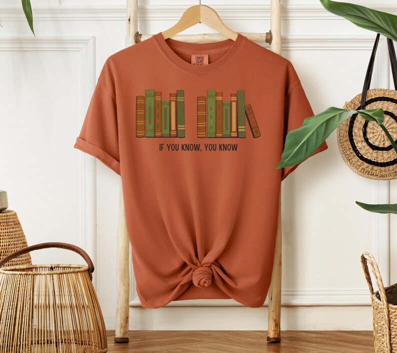 May include: Rust-colored t-shirt featuring a graphic of a bookshelf with green, brown, and tan books. The text "IF YOU KNOW, YOU KNOW" is printed below the bookshelf. The shirt is hanging on a wooden rack.