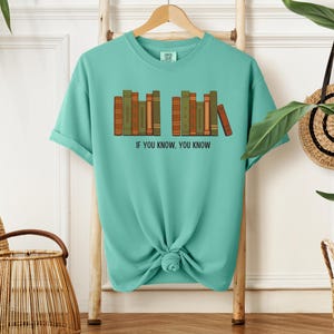 May include: A turquoise t-shirt with a graphic of a row of books in shades of brown and green. The text "IF YOU KNOW, YOU KNOW" is printed below the books. The shirt is hanging on a wooden rack.