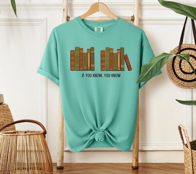 May include: A turquoise t-shirt with a graphic of a row of books in shades of brown and green. The text "IF YOU KNOW, YOU KNOW" is printed below the books. The shirt is hanging on a wooden rack.