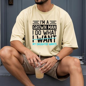 May include: A light yellow t-shirt with the text "I'M A GROWN MAN I DO WHAT I WANT MY DAUGHTER WANTS" in black and blue. The shirt is worn by a person sitting down, wearing a watch and holding a drink.