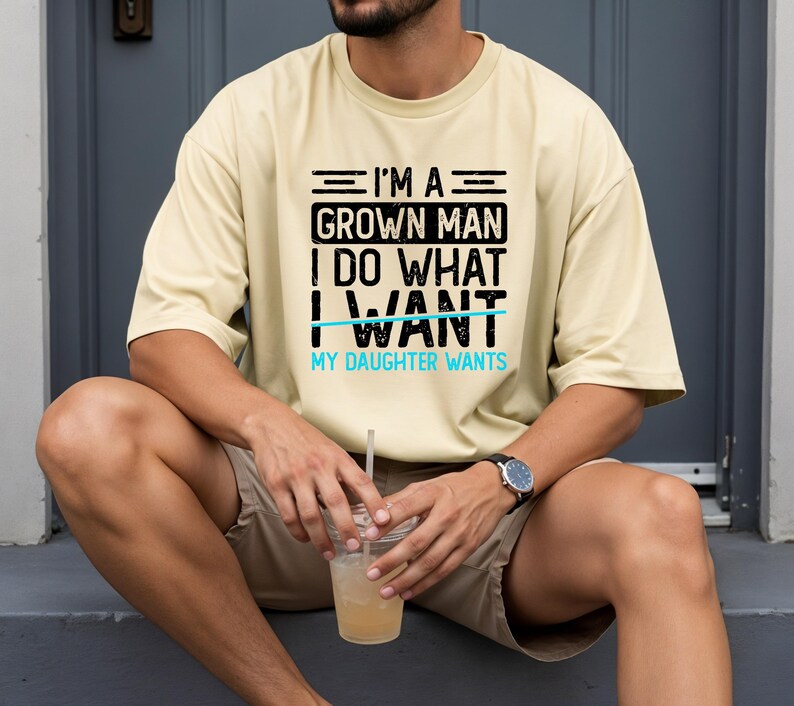 May include: A light yellow t-shirt with the text "I'M A GROWN MAN I DO WHAT I WANT MY DAUGHTER WANTS" in black and blue. The shirt is worn by a person sitting down, wearing a watch and holding a drink.