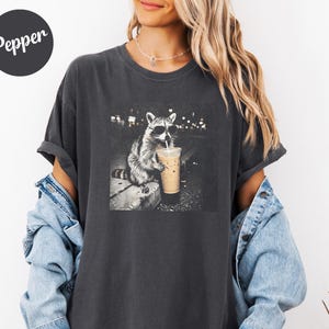 May include: Dark gray t-shirt featuring a black and white image of a raccoon wearing sunglasses and drinking an iced coffee. The raccoon is sitting on a ledge with a city background. The word "Pepper" is on the top left of the shirt.