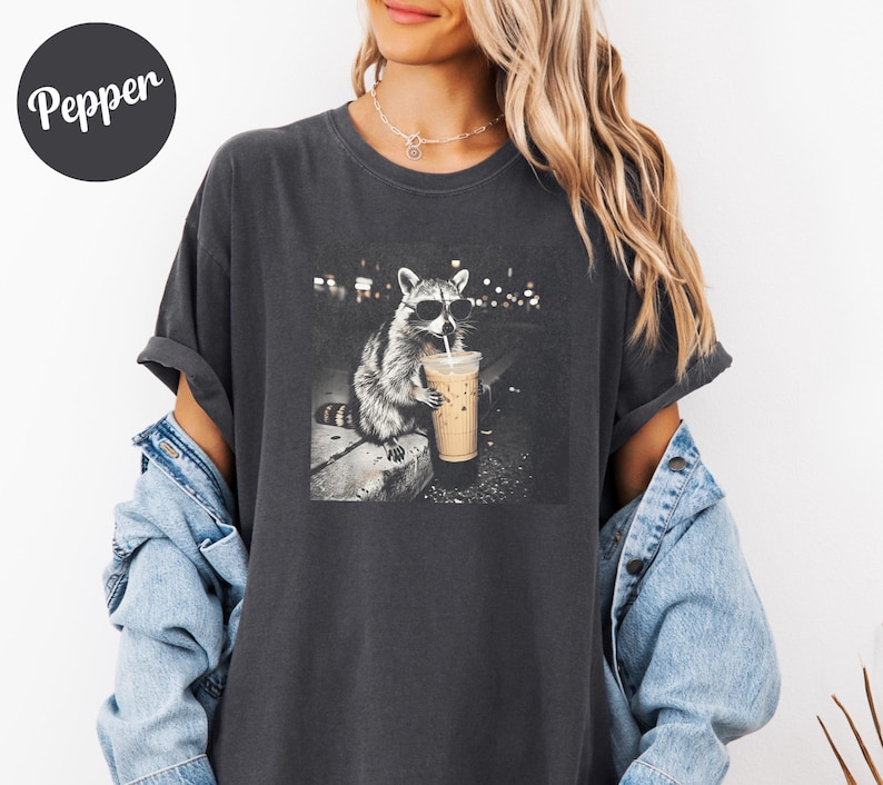 May include: Dark gray t-shirt featuring a black and white image of a raccoon wearing sunglasses and drinking an iced coffee. The raccoon is sitting on a ledge with a city background. The word "Pepper" is on the top left of the shirt.