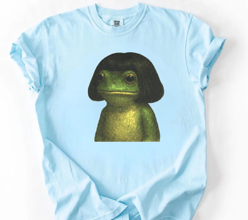 May include: Light blue t-shirt featuring a whimsical illustration of a green frog wearing a black wig. The frog has large, expressive eyes and a detailed, textured appearance. The shirt is made of a soft material.
