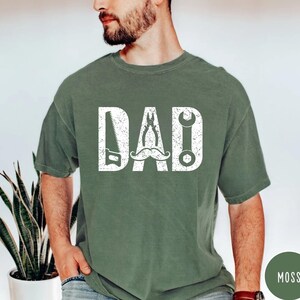 May include: A moss green t-shirt with the word "DAD" in white, incorporating a saw, pliers, a moustache, and a spanner. The shirt is a solid colour and has a crew neck.