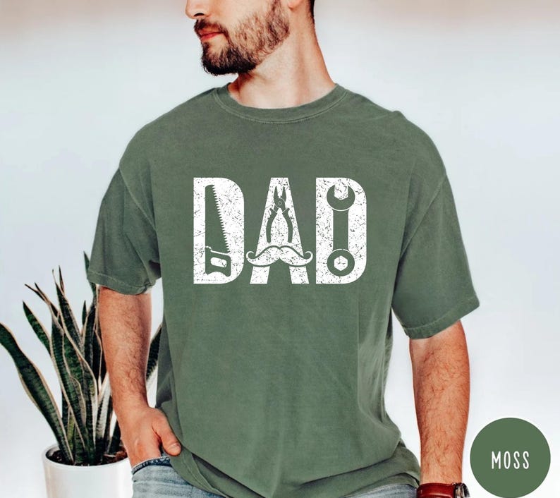 May include: A moss green t-shirt with the word "DAD" in white, incorporating a saw, pliers, a moustache, and a spanner. The shirt is a solid colour and has a crew neck.
