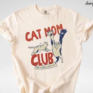 May include: Ivory-colored t-shirt with a red and blue graphic. The graphic reads "CAT MOM CLUB" with a cat illustration. The text "life is better with cats" is below the club text. The shirt has short sleeves and a crew neck.