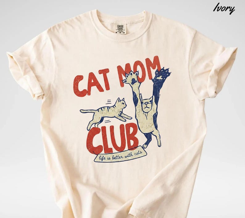 May include: Ivory-colored t-shirt with a red and blue graphic. The graphic reads "CAT MOM CLUB" with a cat illustration. The text "life is better with cats" is below the club text. The shirt has short sleeves and a crew neck.