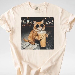 May include: Cream-colored t-shirt featuring a graphic of an orange cat wearing sunglasses and drinking iced coffee. The cat is sitting on a ledge with a blurred city background. The t-shirt has a classic crew neck and short sleeves.