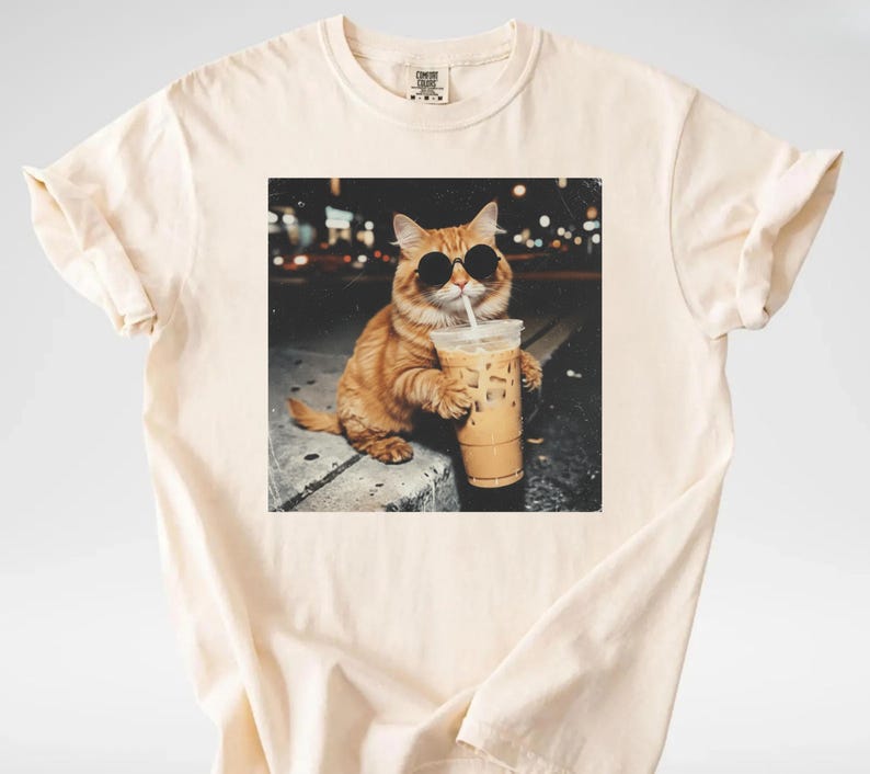 May include: Cream-colored t-shirt featuring a graphic of an orange cat wearing sunglasses and drinking iced coffee. The cat is sitting on a ledge with a blurred city background. The t-shirt has a classic crew neck and short sleeves.