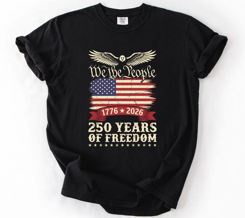 May include: Black t-shirt with a patriotic design. The design includes an American flag, the text "We the People," and "250 YEARS OF FREEDOM" with the years 1776 and 2026. The design also includes an eagle with outstretched wings.