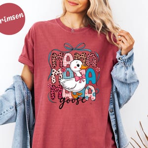 May include: Crimson-colored t-shirt featuring a graphic of a white cartoon goose with a pink bow, surrounded by the words "Mama Goose" in various patterned fonts. The design includes floral accents and a blue bow.