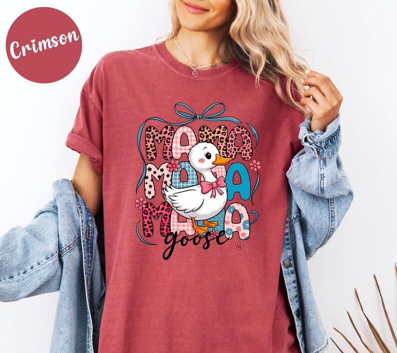 May include: Crimson-colored t-shirt featuring a graphic of a white cartoon goose with a pink bow, surrounded by the words "Mama Goose" in various patterned fonts. The design includes floral accents and a blue bow.