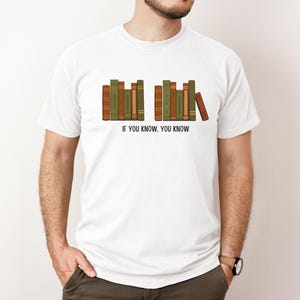 May include: White t-shirt featuring a graphic of a row of books in shades of green and brown. The text "IF YOU KNOW, YOU KNOW" is printed below the books. The shirt is a casual style.