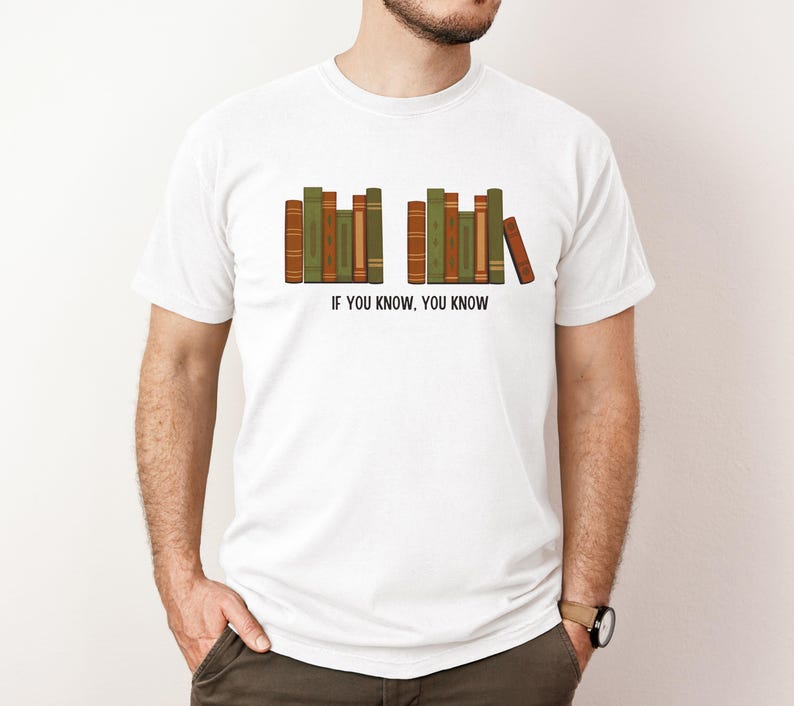May include: White t-shirt featuring a graphic of a row of books in shades of green and brown. The text "IF YOU KNOW, YOU KNOW" is printed below the books. The shirt is a casual style.