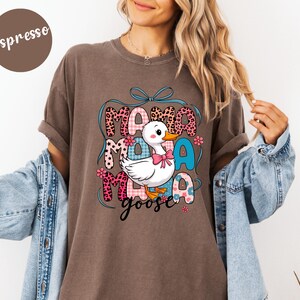 May include: Espresso-colored t-shirt with a graphic of a white duck wearing a pink bow, the words "Mama Goose" in a mix of fonts and patterns. The design includes floral accents and a blue ribbon.