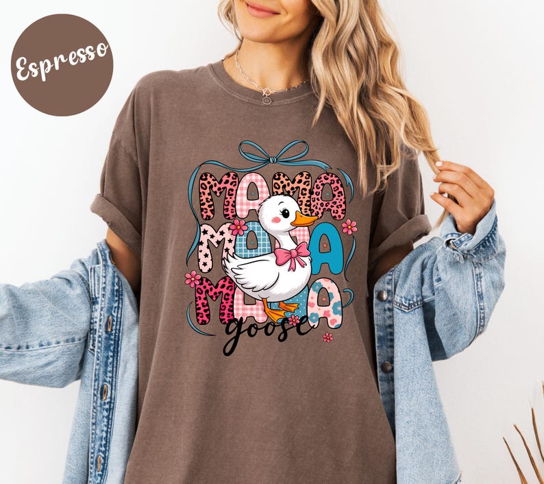 May include: Espresso-colored t-shirt with a graphic of a white duck wearing a pink bow, the words "Mama Goose" in a mix of fonts and patterns. The design includes floral accents and a blue ribbon.