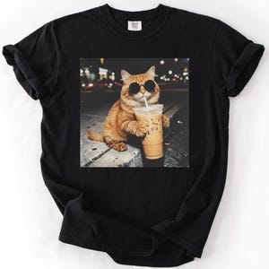 May include: Black t-shirt featuring a graphic of an orange cat wearing sunglasses and holding an iced coffee. The cat is sitting on a ledge with a blurred city background. The image is printed on a black, short-sleeved t-shirt.
