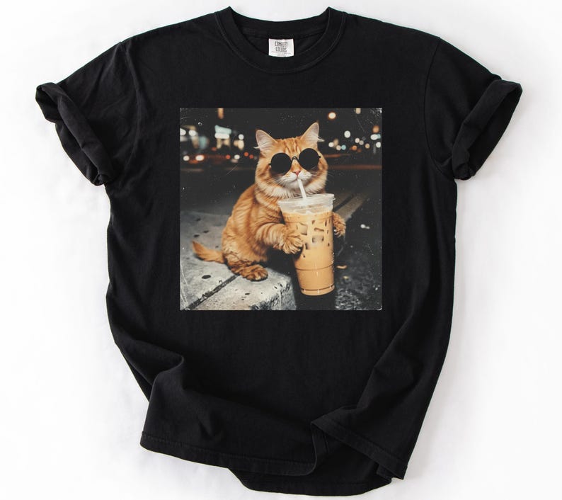 May include: Black t-shirt featuring a graphic of an orange cat wearing sunglasses and holding an iced coffee. The cat is sitting on a ledge with a blurred city background. The image is printed on a black, short-sleeved t-shirt.