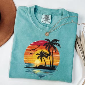 May include: A turquoise Comfort Colors t-shirt with an embroidered sunset design featuring palm trees, birds, and a vibrant orange, yellow, and red sky. A gold necklace with a pendant rests on the shirt. The shirt is size S-P-CH.