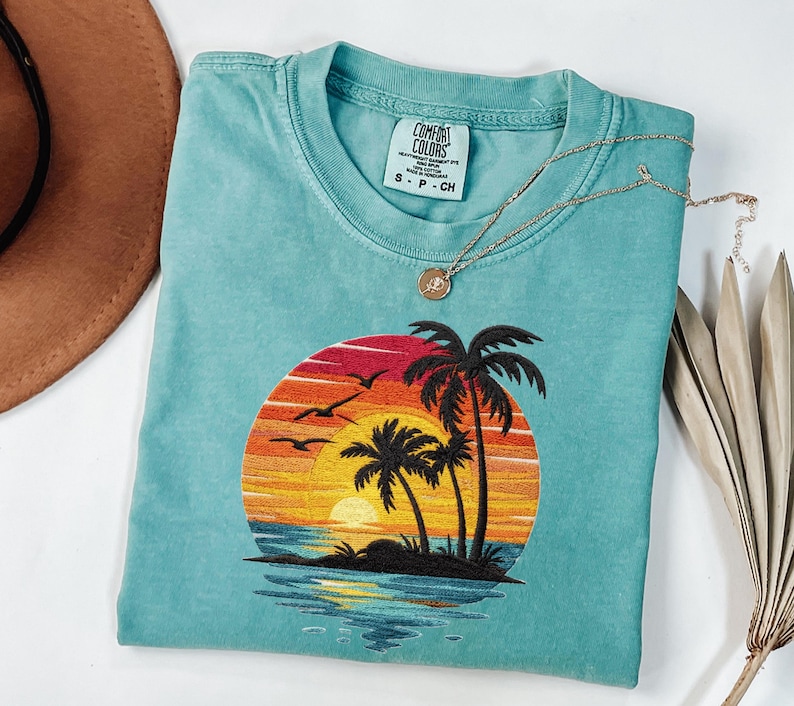 May include: A turquoise Comfort Colors t-shirt with an embroidered sunset design featuring palm trees, birds, and a vibrant orange, yellow, and red sky. A gold necklace with a pendant rests on the shirt. The shirt is size S-P-CH.