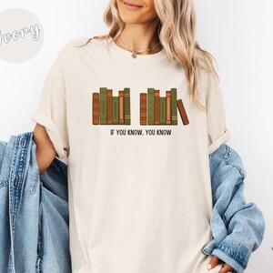 May include: Ivory-colored t-shirt featuring a graphic of stacked books in shades of green and brown. The text "IF YOU KNOW, YOU KNOW" is printed below the books. The shirt has a relaxed fit.