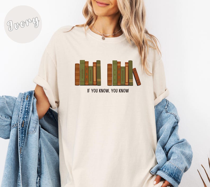 May include: Ivory-colored t-shirt featuring a graphic of stacked books in shades of green and brown. The text "IF YOU KNOW, YOU KNOW" is printed below the books. The shirt has a relaxed fit.