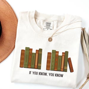 May include: Cream-colored t-shirt with a graphic of stacked books in shades of brown and green. The text "IF YOU KNOW, YOU KNOW" is printed below the books. A gold necklace and a brown hat are also in the image.