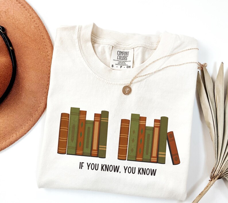 May include: Cream-colored t-shirt with a graphic of stacked books in shades of brown and green. The text "IF YOU KNOW, YOU KNOW" is printed below the books. A gold necklace and a brown hat are also in the image.