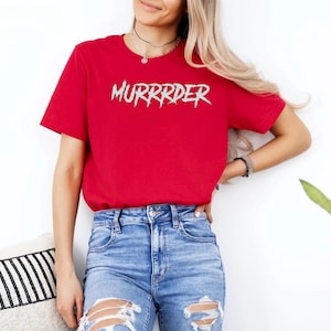 May include: A red t-shirt with the word "MURRRDER" in a spiky, silver font. The shirt is being worn with blue jeans. The person is standing in front of a white wall.